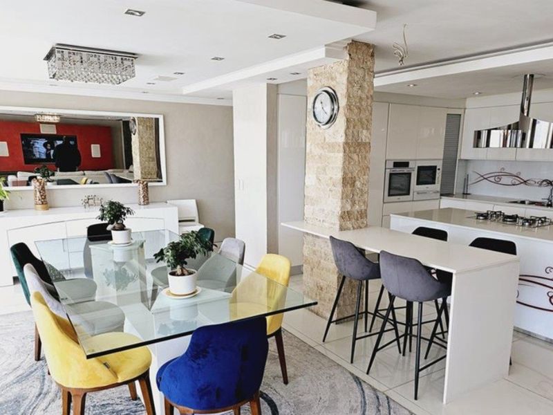 Modern open-plan living and kitchen area featuring a stone-clad pillar, sleek gloss cabinetry, integrated appliances, and a breakfast bar.