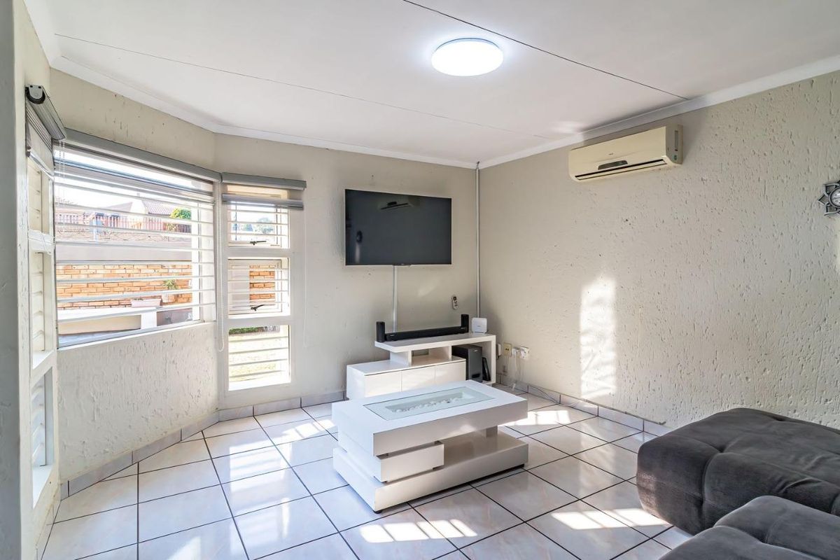 A bright living room featuring tiled flooring, a wall-mounted air conditioning unit, and large windows with aluminium frames.