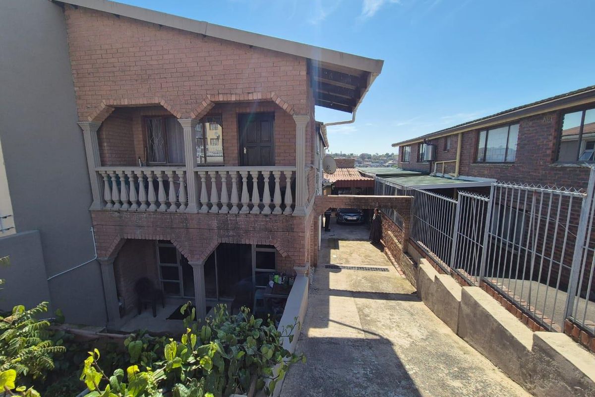 Rear view of a multi-level face brick house featuring a balcony with balustrades, a concrete driveway, and a covered carport area.