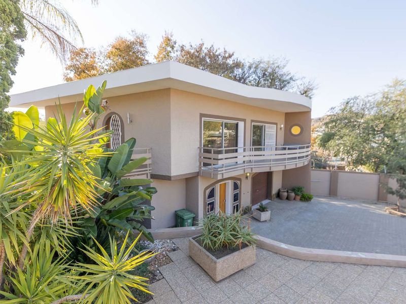 A two-story residential property featuring a curved balcony, paved driveway, and integrated garage, set within a landscaped garden environment.