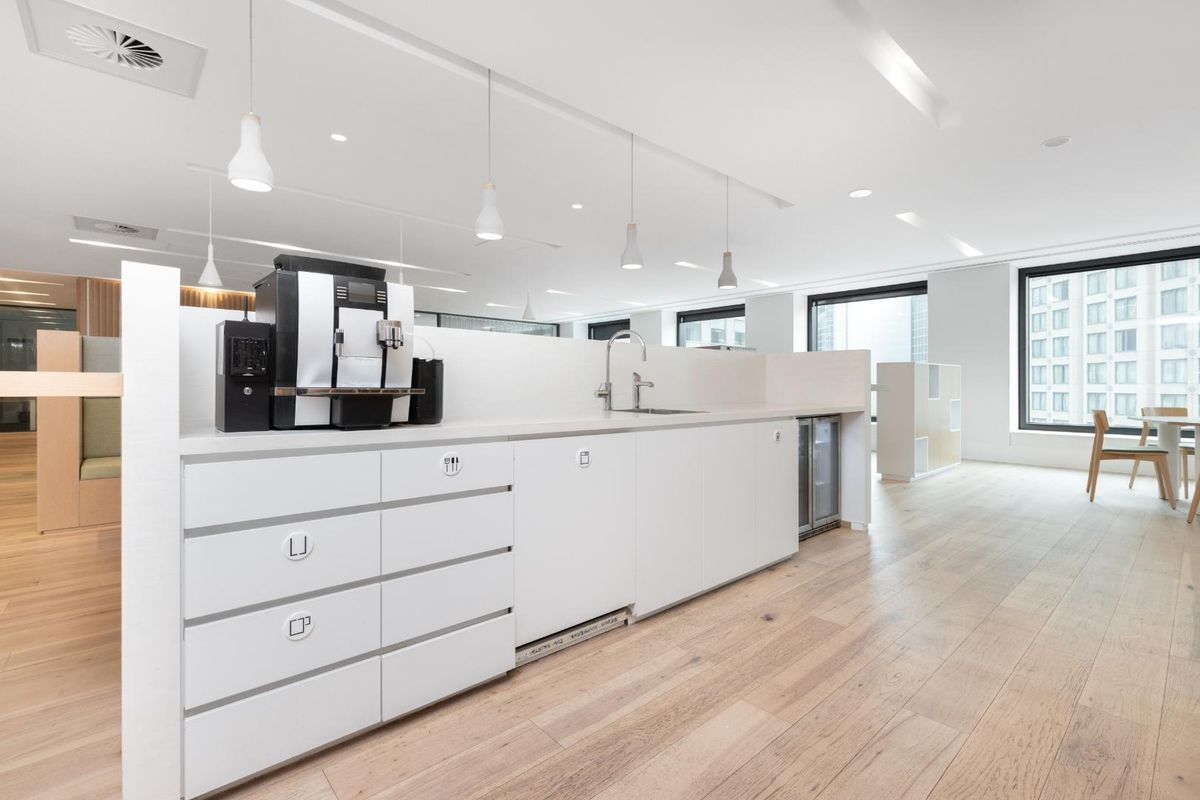 Modern office kitchenette featuring sleek white cabinetry, integrated sink, and minimalist pendant lighting, set within a bright, open-plan workspace with timber flooring.