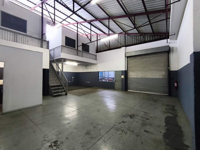 Spacious industrial warehouse unit featuring a roller shutter door, mezzanine level with staircase, and high exposed steel roof trusses.