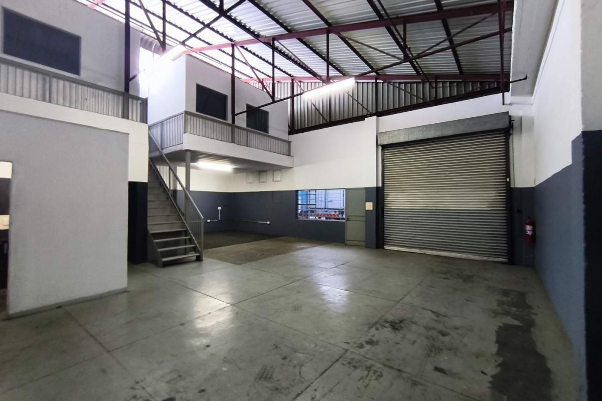 Spacious industrial warehouse unit featuring a roller shutter door, mezzanine level with staircase, and high exposed steel roof trusses.