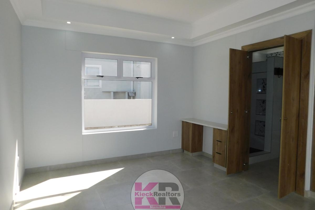 A bright bedroom featuring neutral tiled flooring, a built-in desk unit, recessed ceiling lighting, and an en-suite bathroom entrance.