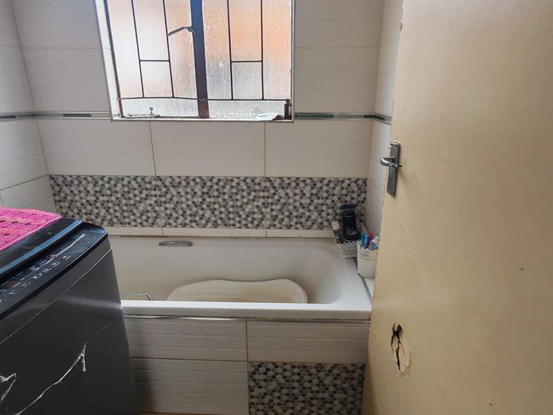 A compact bathroom featuring a built-in bathtub, tiled walls with decorative accents, and a window with security bars. The door requires repair.