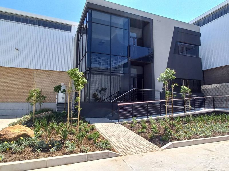 Modern industrial office entrance featuring a striking glass facade, contemporary metal cladding, and a well-maintained landscaped approach with paved pedestrian access.