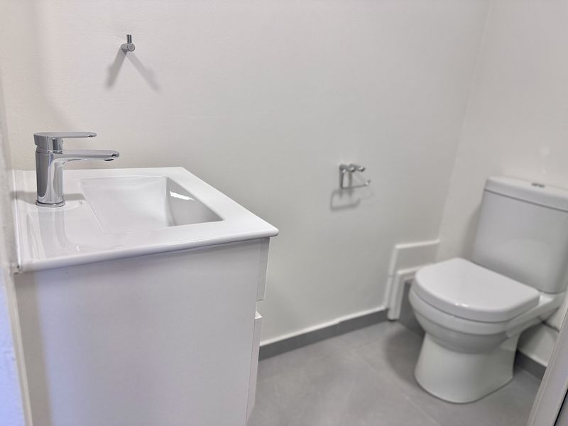 A clean, modern bathroom featuring a white vanity with basin, chrome faucet, and a standard toilet with grey tiled flooring.