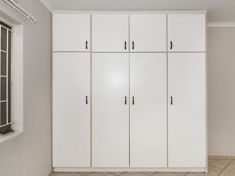 A clean bedroom featuring floor-to-ceiling built-in cupboards, tiled flooring, and a window fitted with security bars.