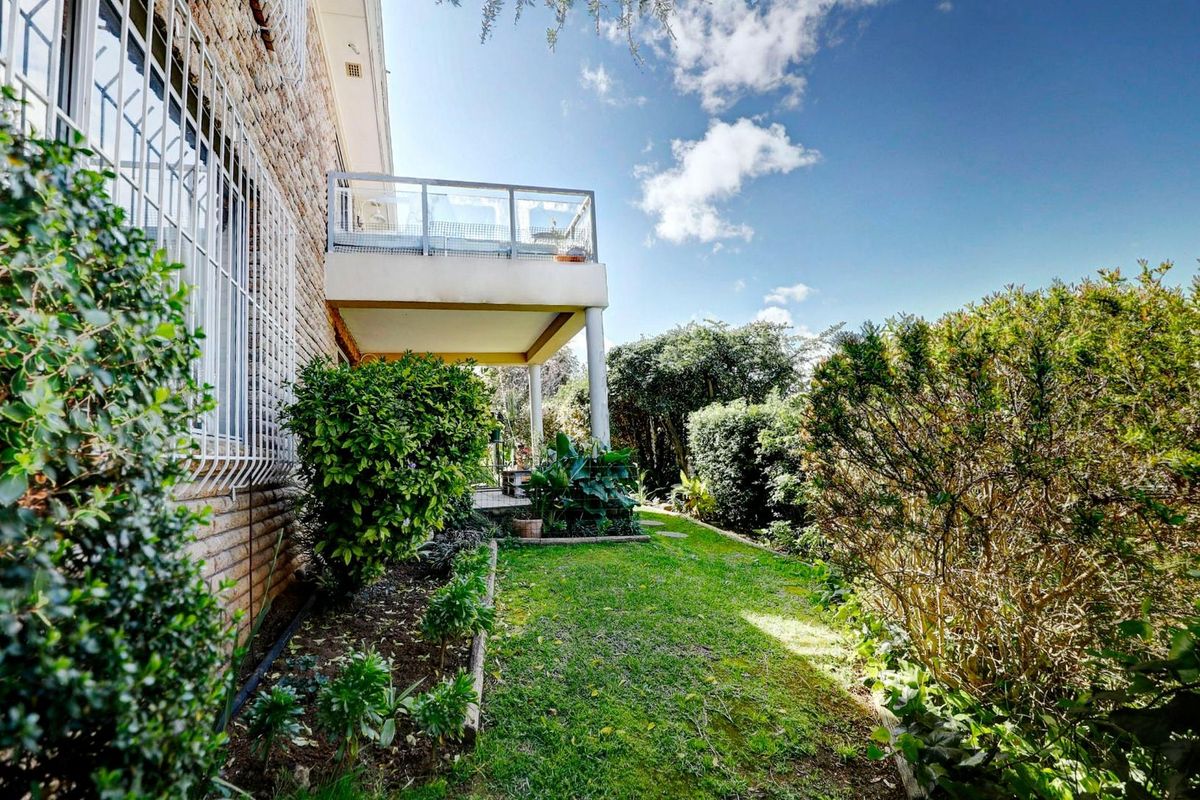 A view of the rear garden featuring a raised balcony with glass balustrades and established landscaping along the brick townhouse exterior.