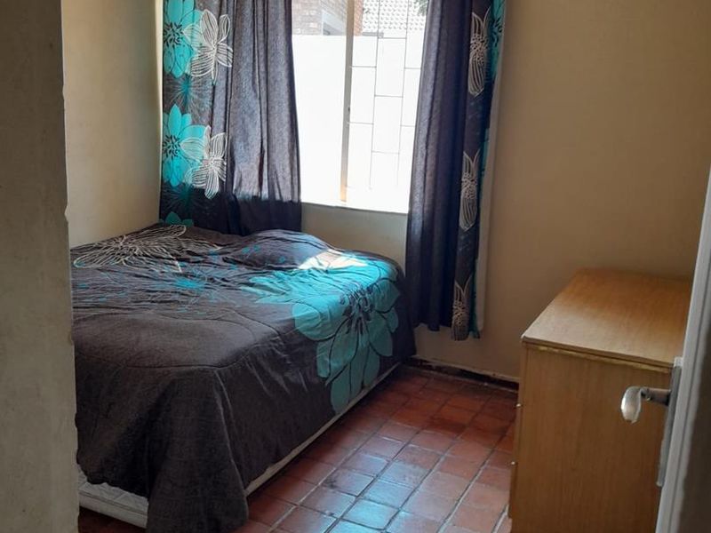 A compact bedroom featuring terracotta tiled flooring, a ceiling fan, and a window fitted with security bars.