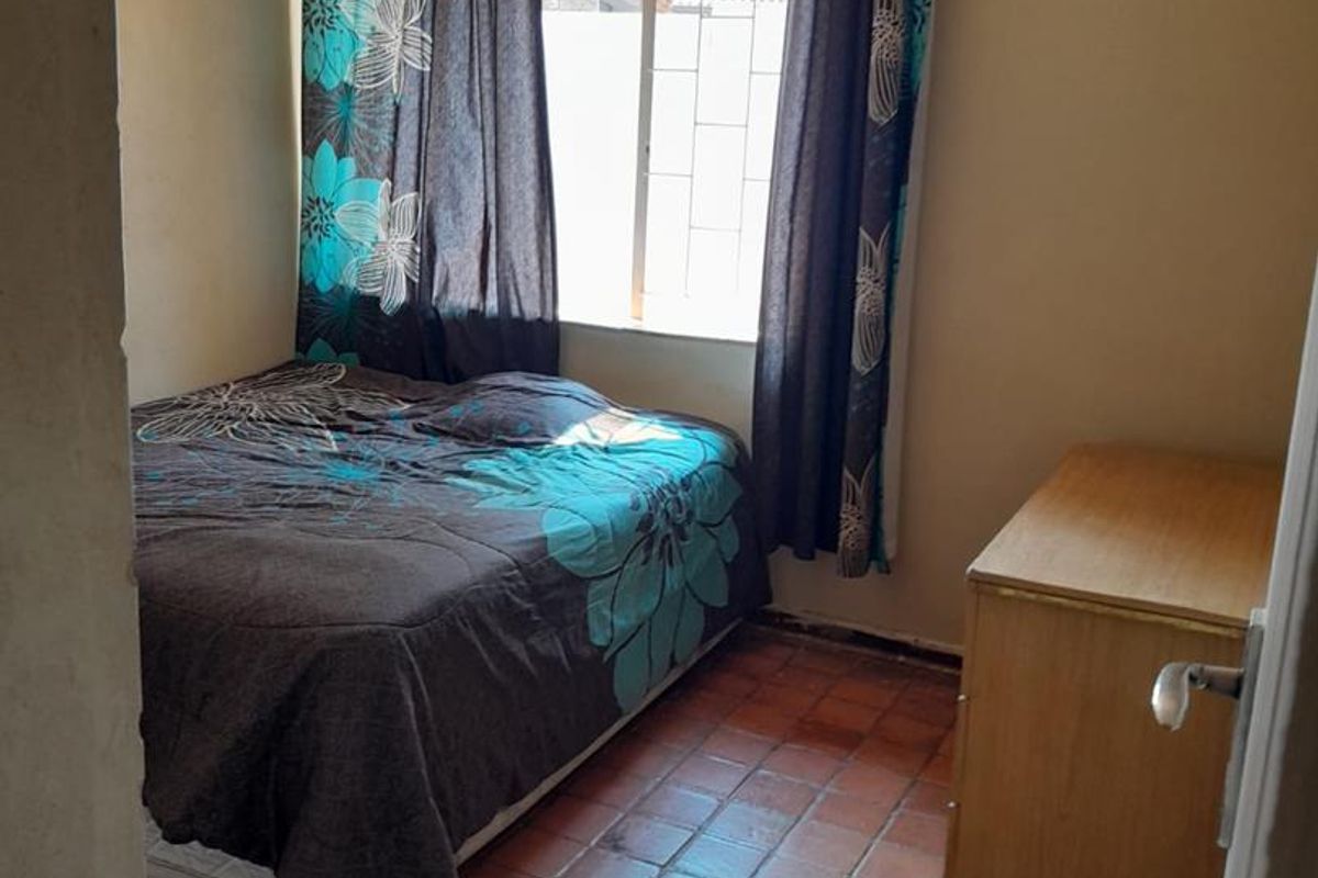A compact bedroom featuring terracotta tiled flooring, a ceiling fan, and a window fitted with security bars.