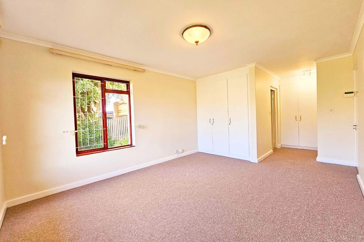 A spacious, carpeted bedroom featuring built-in cupboards, a ceiling light fixture, and a window with security bars.