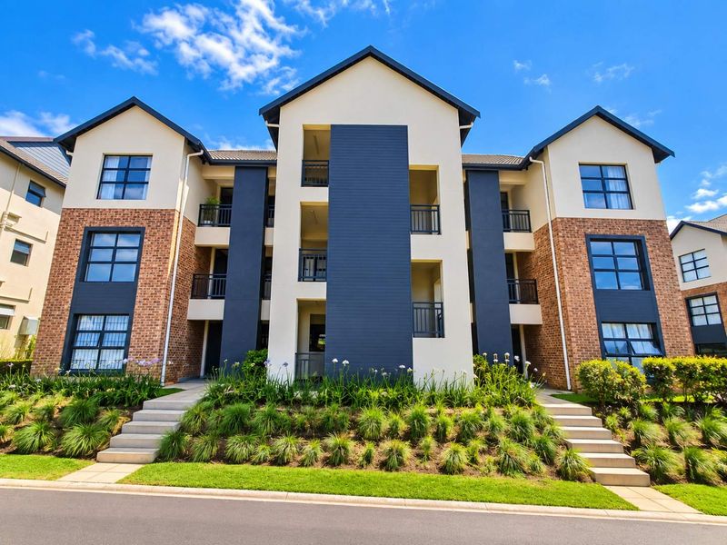 Front elevation of a modern multi-story apartment building featuring brick and painted facade, private balconies with metal railings, and landscaped garden beds.