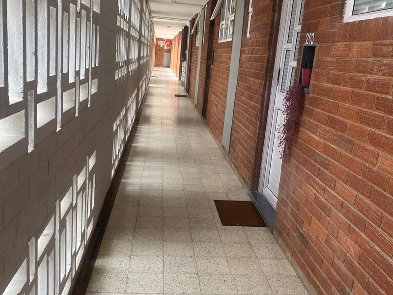 A long, open-air apartment hallway featuring exposed brick walls, decorative breeze block screening, and tiled flooring.
