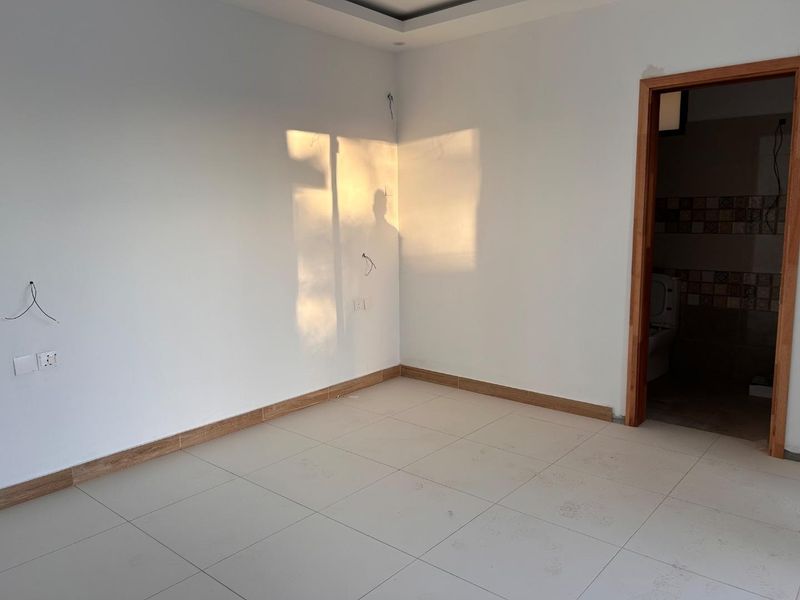 An empty bedroom featuring tiled flooring, recessed ceiling detail, and an adjoining bathroom, currently awaiting final electrical finishes and personal touches.