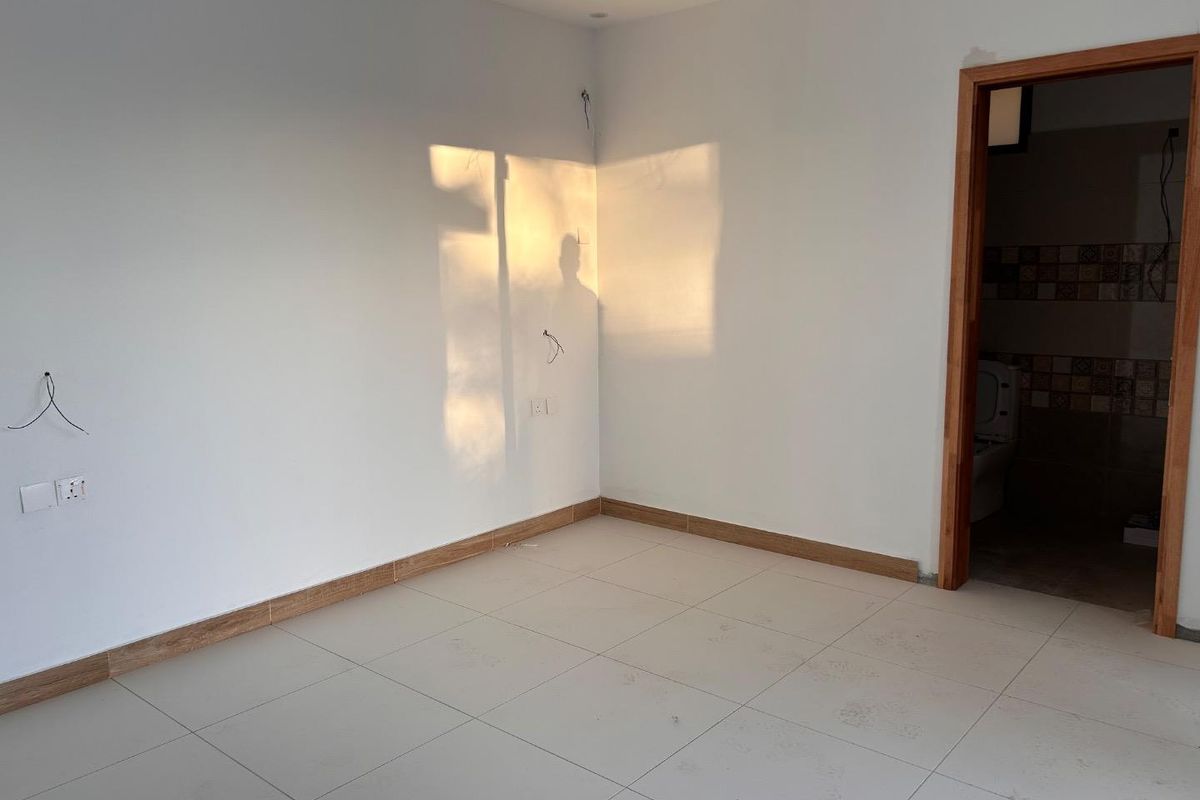 An empty bedroom featuring tiled flooring, recessed ceiling detail, and an adjoining bathroom, currently awaiting final electrical finishes and personal touches.