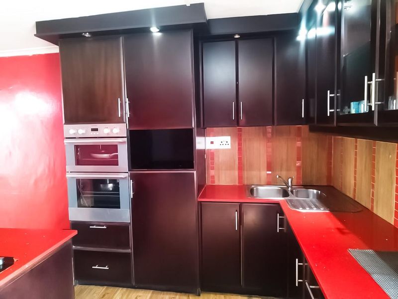 A functional kitchen featuring dark wood cabinetry, a built-in oven and microwave, red countertops, and a double sink with tiled backsplash.