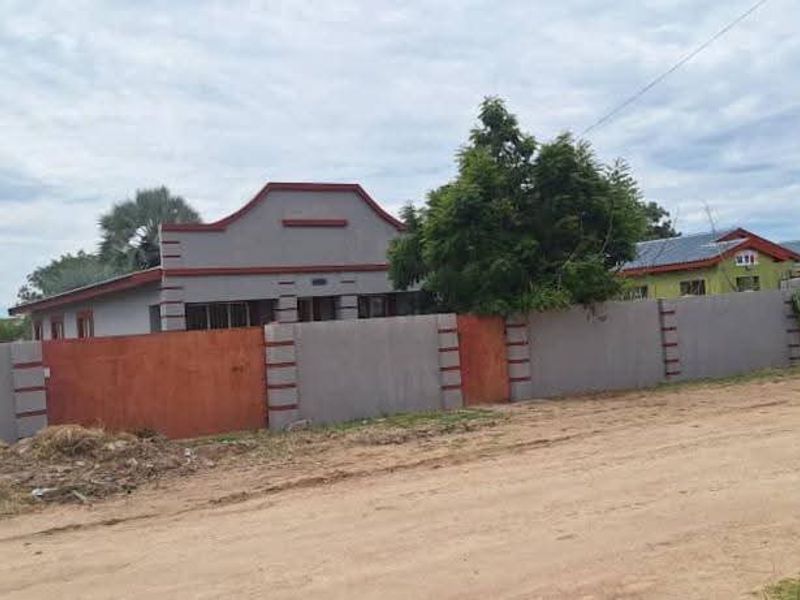 Front view of a residential property featuring a gabled roof and a substantial perimeter wall, situated along an unpaved road.