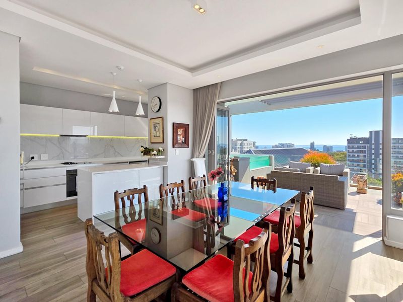 Modern open-plan kitchen and dining area featuring sleek white cabinetry, marble-look finishes, and seamless flow to a spacious balcony with city and sea views.