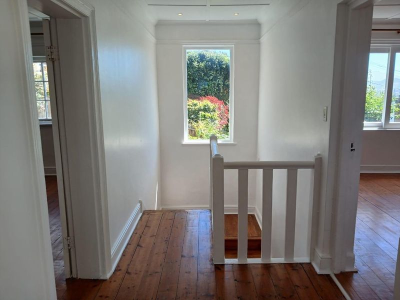 A bright hallway featuring polished timber flooring, a white staircase balustrade, and a large window providing natural light and garden views.