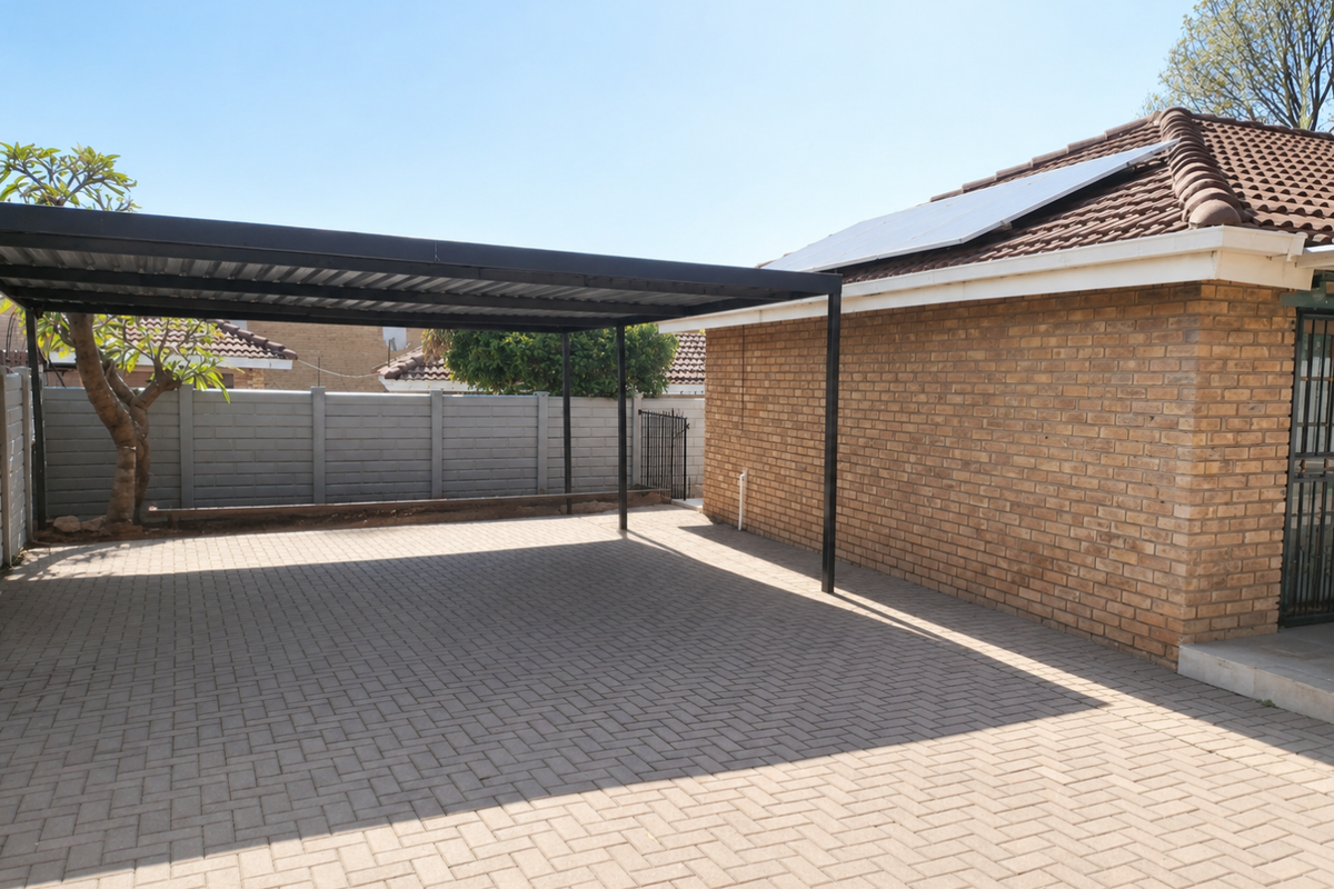 A paved outdoor area featuring a modern black metal carport structure, brick exterior wall, and roof-mounted solar panels.