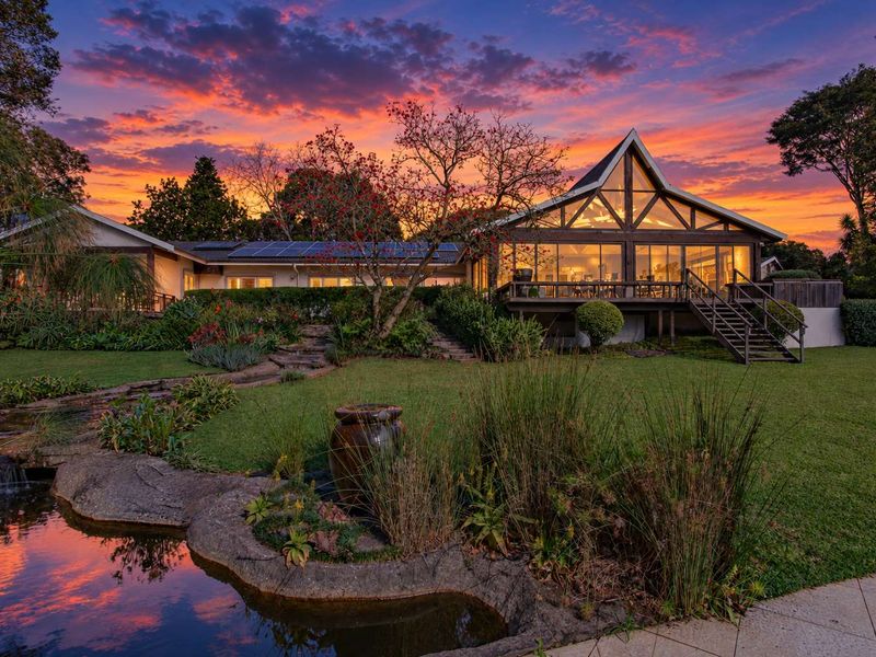 Stunning rear view of a modern home featuring a timber deck, large glass windows, solar panels, and a beautifully landscaped garden with a pond.