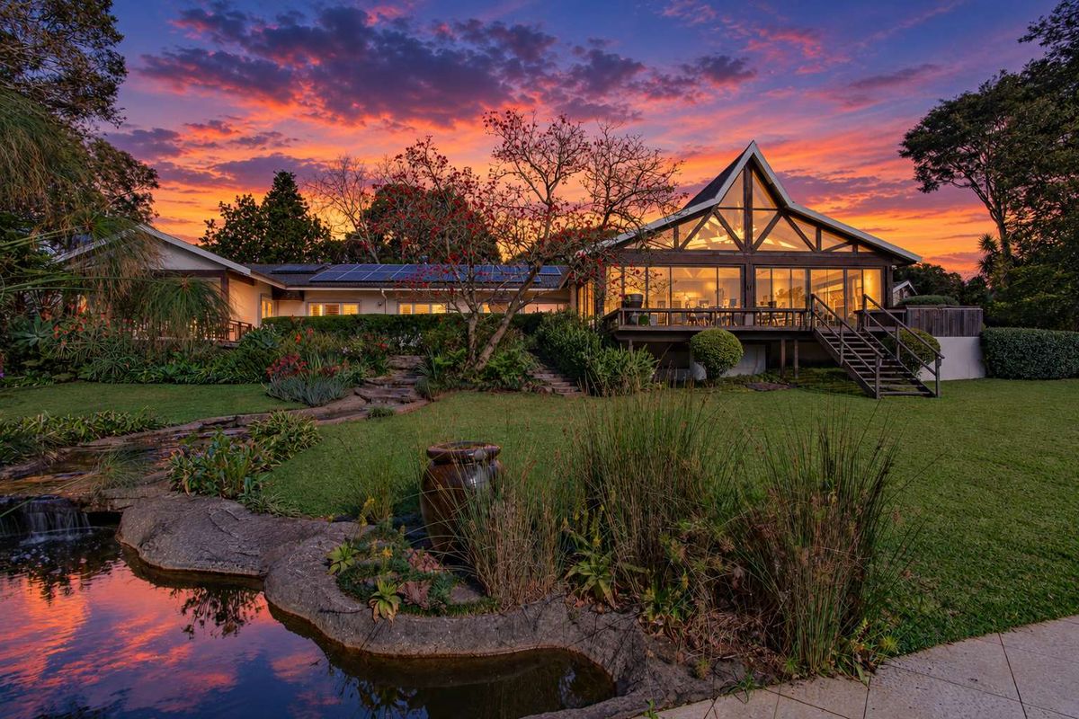 Stunning rear view of a modern home featuring a timber deck, large glass windows, solar panels, and a beautifully landscaped garden with a pond.