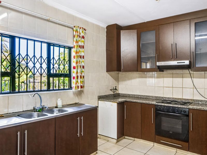 Functional kitchen featuring dark wood cabinetry, granite countertops, built-in oven, hob, extractor fan, and a double stainless steel sink with security window bars.