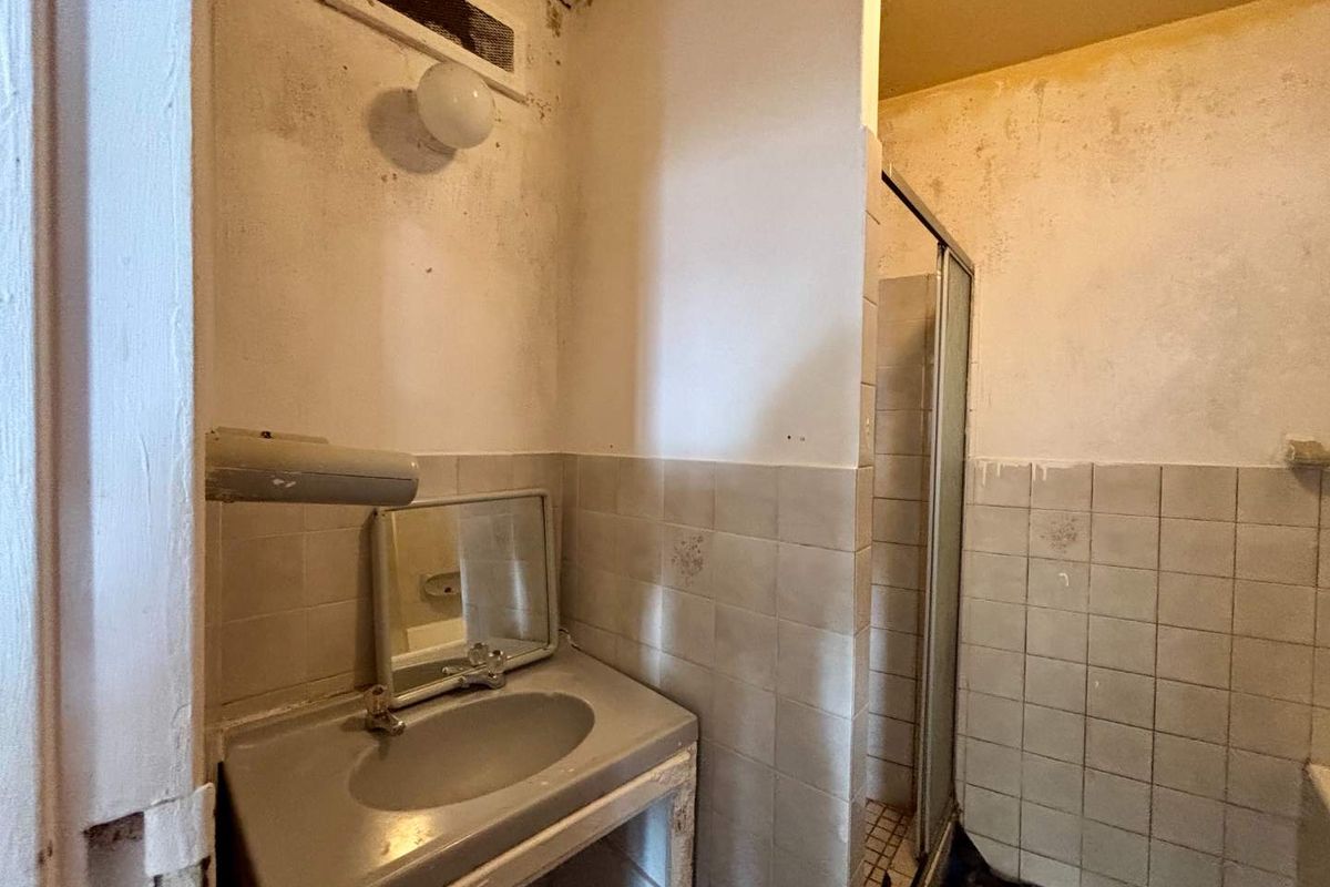 A dated bathroom requiring significant renovation, featuring a built-in vanity basin and a shower enclosure with visible wear and wall surface deterioration.