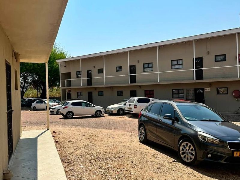 Exterior view of an apartment complex featuring a tiled walkway, metal balcony railings, and a gravel parking area.