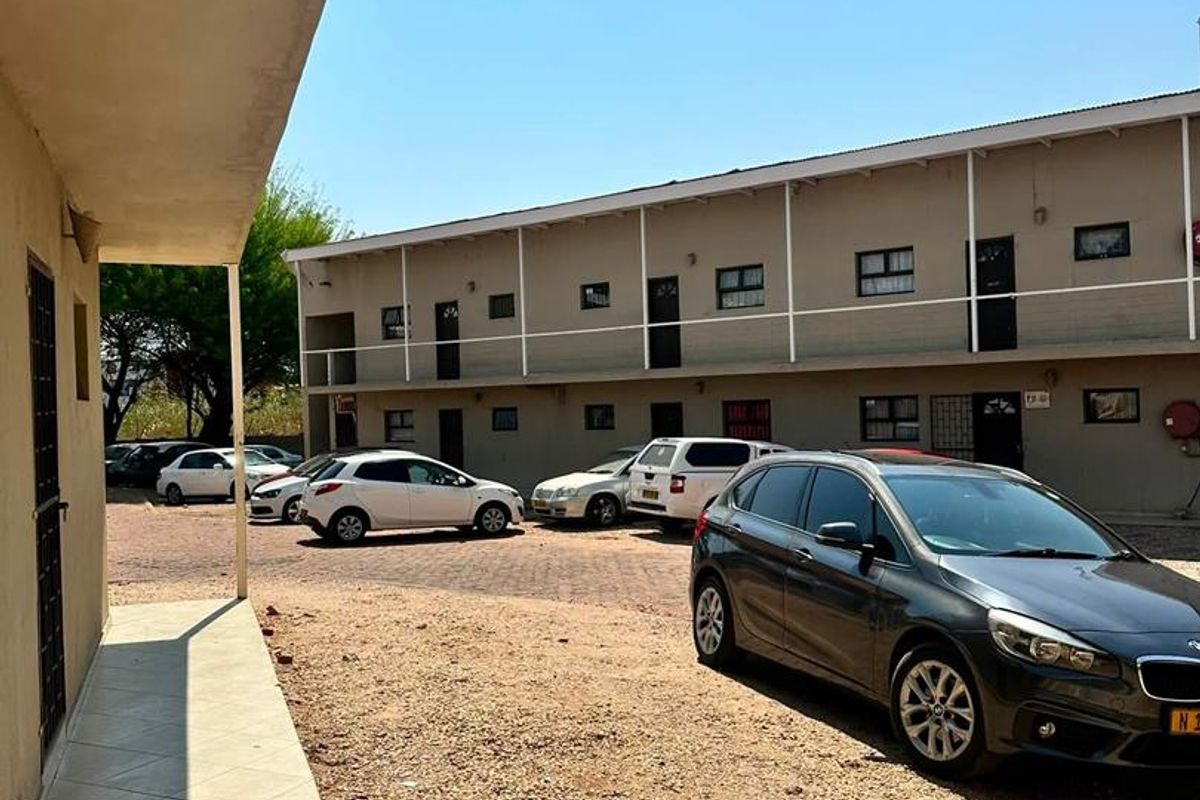 Exterior view of an apartment complex featuring a tiled walkway, metal balcony railings, and a gravel parking area.