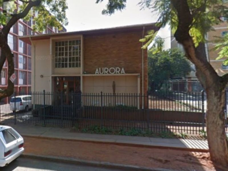 Street-level view of the Aurora apartment building facade, featuring brick exterior, metal security fencing, and established trees.
