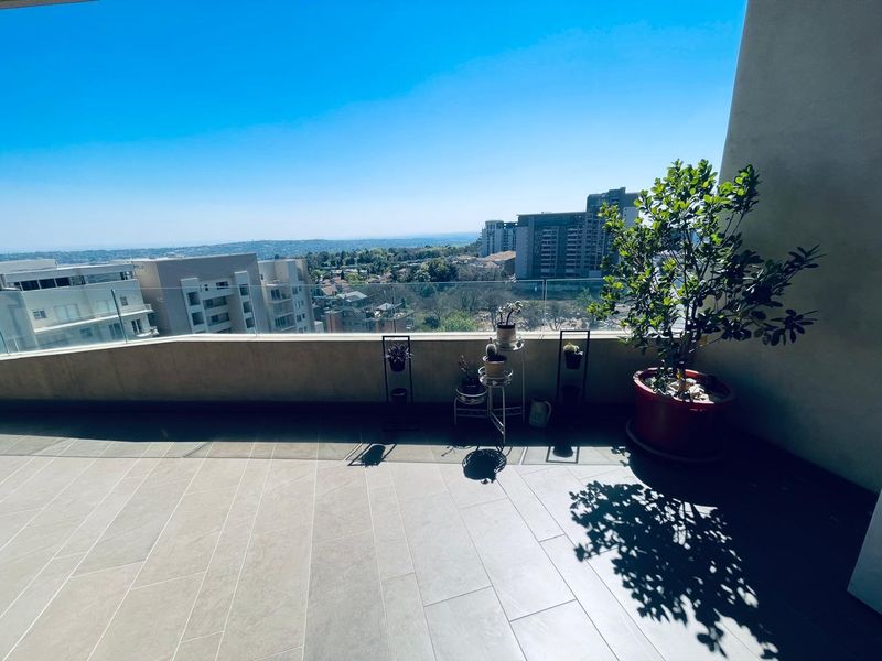 A spacious, modern balcony featuring sleek tiled flooring and a clear glass balustrade, offering expansive elevated views of the surrounding urban landscape.