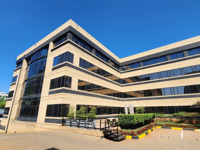 A modern multi-story office building featuring a structured facade with extensive tinted glass windows, paved parking area, and manicured garden beds.