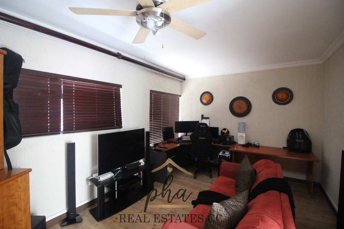 A living room featuring a ceiling fan, wooden blinds, and laminate flooring, currently utilized as a multi-purpose living and workspace area.