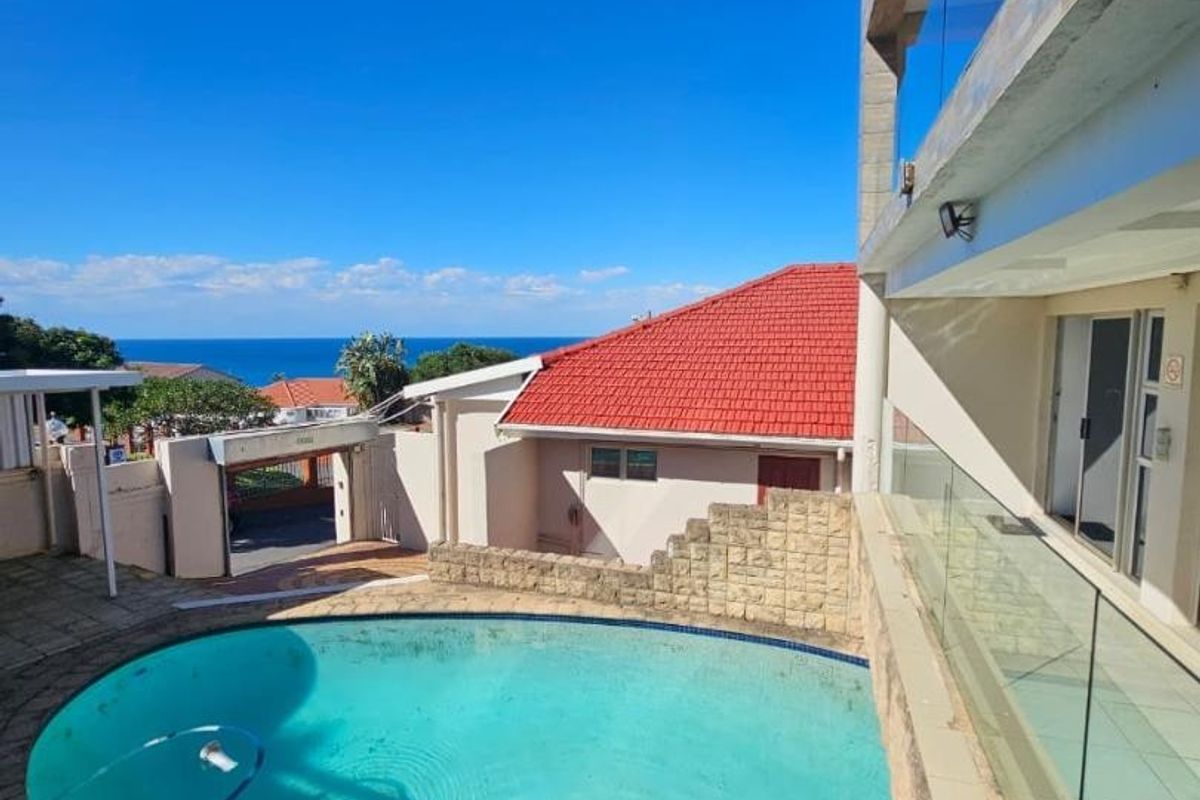 Outdoor pool area featuring a paved deck, glass balustrades, and a stone retaining wall with views of the surrounding neighborhood and ocean.