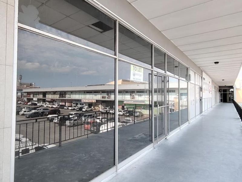 An exterior covered walkway featuring large glass storefront windows, a metal security gate, and concrete flooring, overlooking a parking area.