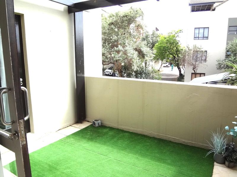 A compact balcony featuring artificial grass, a solid balustrade, and a partial pergola structure, offering an outdoor space accessible via a glass door.