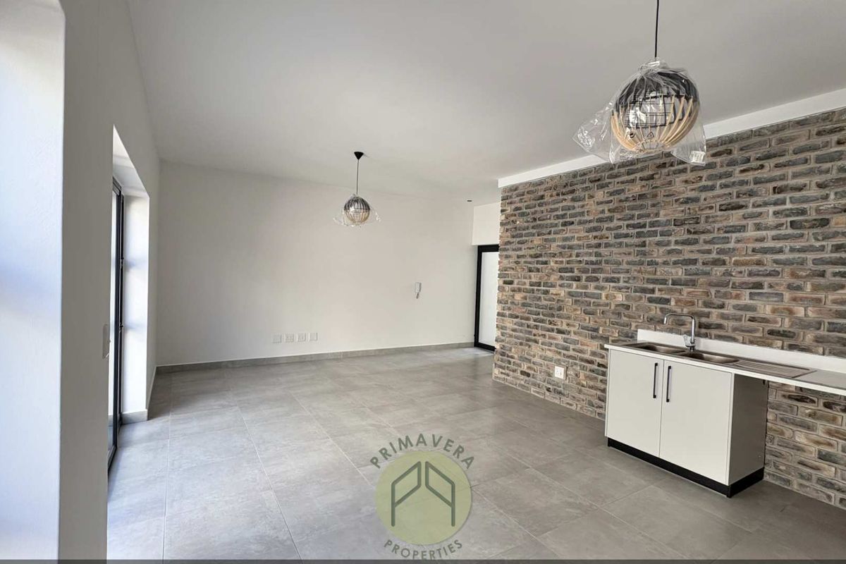 An open-plan living area featuring large format floor tiles, a striking exposed brick feature wall, and a compact kitchenette with a stainless steel sink.