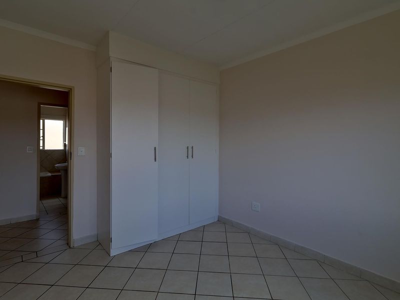 A clean, empty bedroom featuring neutral tiled flooring and white built-in cupboards, with an adjacent view into a bathroom.