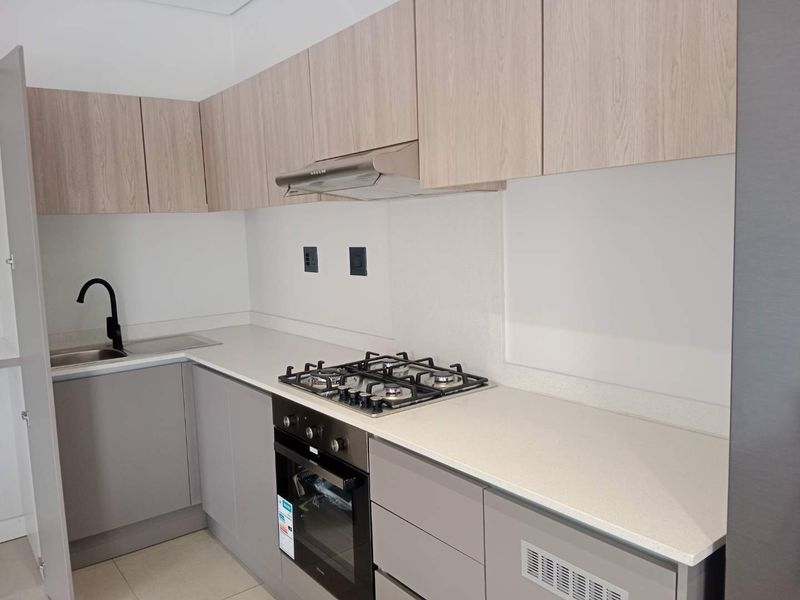 Modern kitchen featuring light wood-tone upper cabinets, grey lower cabinetry, stone countertops, a gas hob, built-in oven, and a sleek black mixer tap.