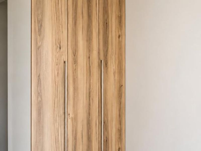 A clean, modern bedroom featuring light-toned wood grain built-in wardrobes, neutral tiled flooring, and a ceiling fan.