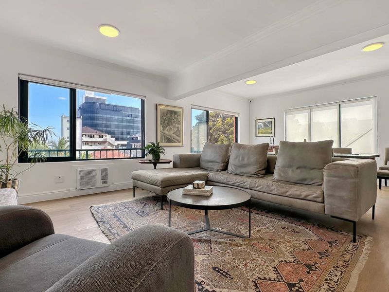 A bright and spacious living room featuring large windows, recessed ceiling lighting, and light-toned laminate flooring with an urban view.