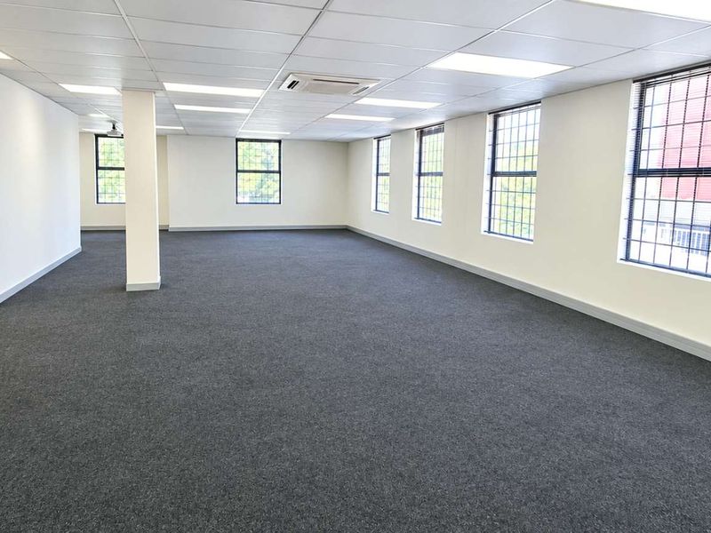 A spacious, open-plan office area featuring neutral carpeting, a suspended ceiling with recessed lighting, and large windows with security bars.