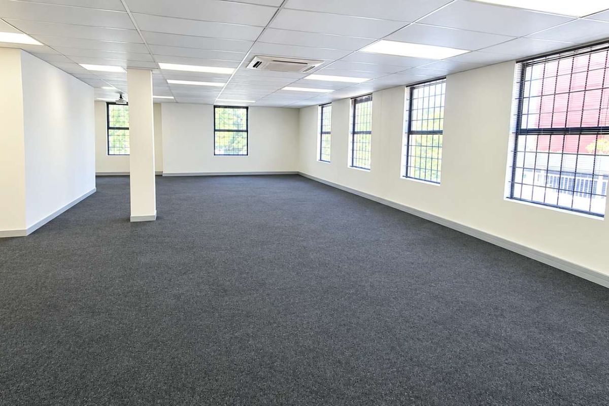 A spacious, open-plan office area featuring neutral carpeting, a suspended ceiling with recessed lighting, and large windows with security bars.