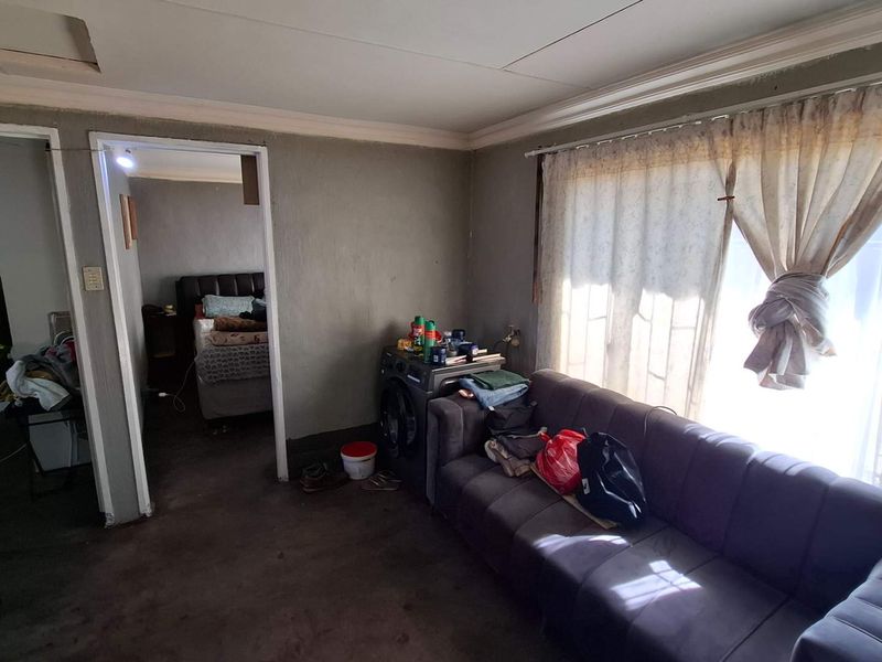 A living room area featuring basic finishes, ceiling cornicing, and window security bars, requiring significant cosmetic updates and repairs to improve its overall condition.