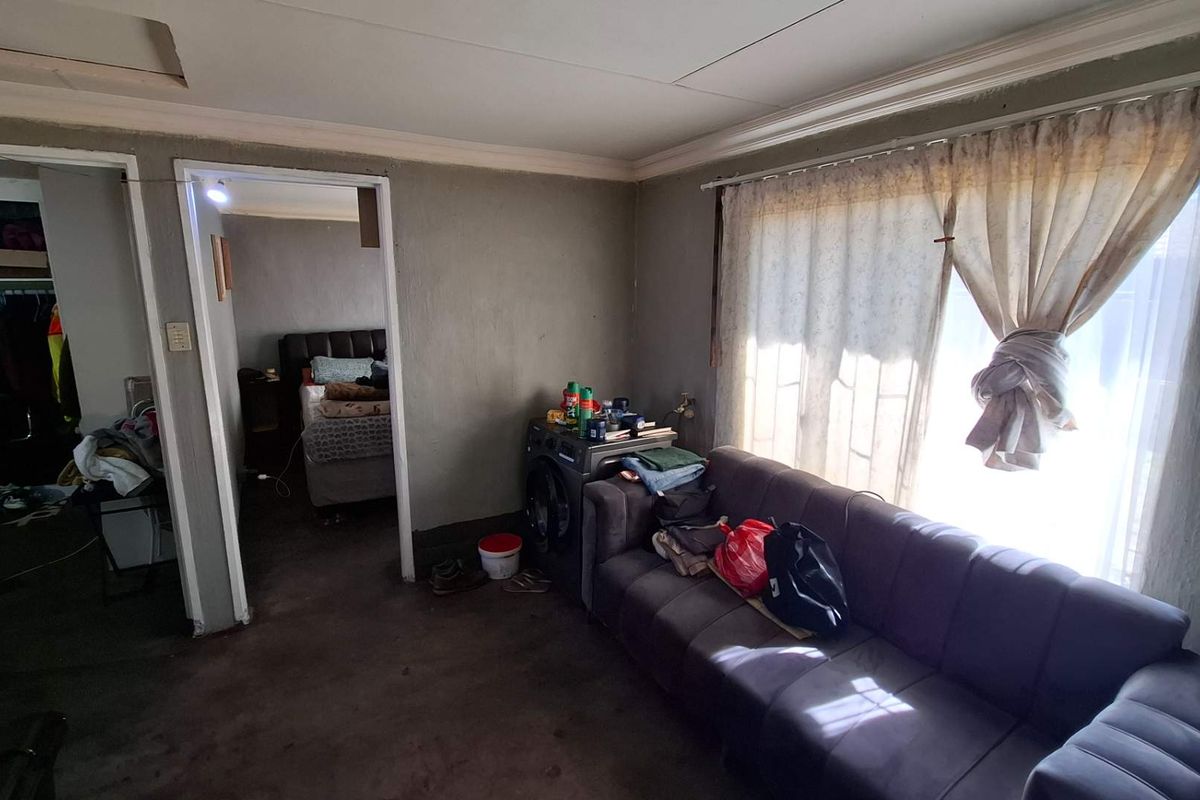 A living room area featuring basic finishes, ceiling cornicing, and window security bars, requiring significant cosmetic updates and repairs to improve its overall condition.
