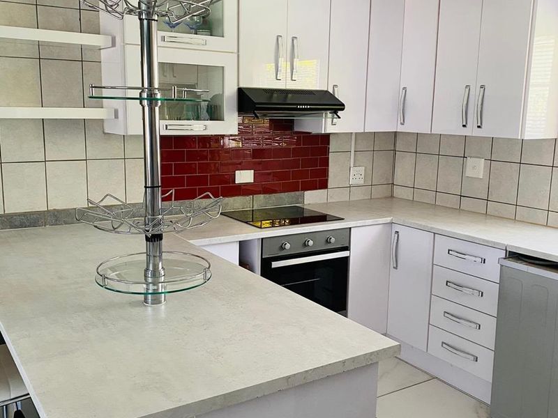 A functional kitchen featuring white cabinetry, contrasting red tiled backsplash, built-in oven, electric hob, and a breakfast bar with stone-look countertops.