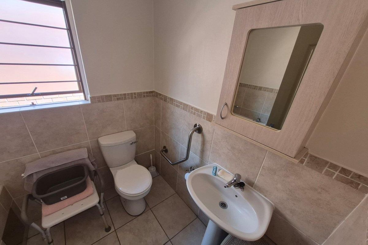 A functional bathroom featuring a pedestal basin, toilet, wall-mounted grab rail, and a mirrored medicine cabinet with neutral wall and floor tiling.