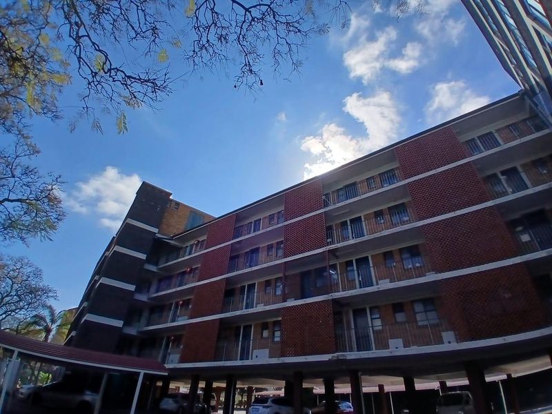 A low-angle view of a multi-story apartment building featuring a brick facade, private balconies with metal railings, and a covered ground-level parking area.